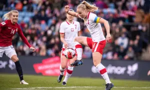 Piłkarka w białej koszulce z czerwonymi szortami kontroluje piłkę. To emocjonujący moment kobiecego futbolu.
