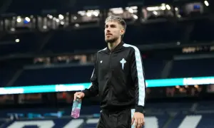 Piłkarz Oscar Mingueza w czarnej bluzie z jasnoniebieskimi pasami, trzymający butelkę napoju izotonicznego na tle stadionu.