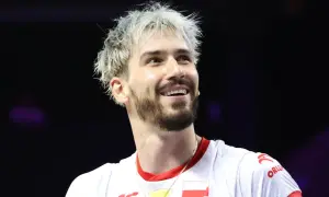 Uśmiechnięty siatkarz z blond włosami, jeden z tych, którzy wygrali Ligę Mistrzów, w białej koszulce z polską flagą.