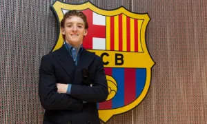Uśmiechnięty Anthony Gordon w garniturze stoi przed herbem FC Barcelony.