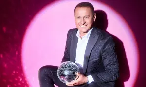 Piotr Świr Świerczewski w eleganckim garniturze trzyma błyszczącą kulę dyskotekową na tle różowego światła.