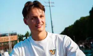Uśmiechnięty kornel lisman w białej koszulce z logo, na tle nieba i zieleni.