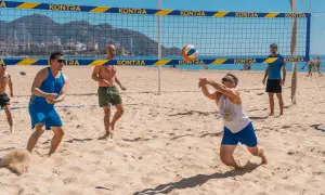 Mężczyźni grają w siatkówkę plażową. Jeden z nich zagrywa piłkę, inni czekają na boisku. To świetny przykład beach soccer Polska!