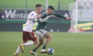 Piłkarze ŁKS i Legii walczą o piłkę na boisku. Jeden z nich w białej koszulce, drugi w zielonej.