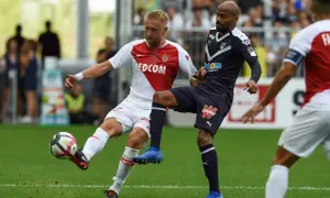 Piłkarz w białej koszulce z herbem **AS Monaco FC** walczy o piłkę z przeciwnikiem w granatowym stroju.
