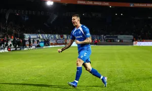 Mateusz Żukowski w niebieskim stroju świętuje bramkę na zielonej murawie stadionu, z trybunami pełnymi kibiców w tle.