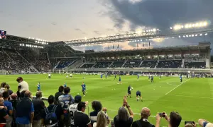 Widownia na stadionie przed meczem FC Bologna. Piłkarze rozgrzewają się na zielonej murawie, a kibice z flagami i aparatami obserwują przygotowania.