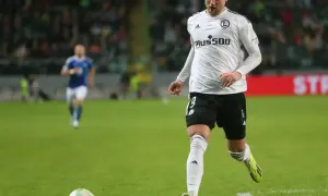 Piłkarz Blaž Kramer w białej koszulce z numerem 19 i czarnych spodenkach biegnie po zielonej murawie, zbliżając się do piłki.