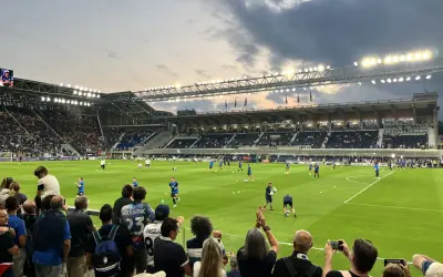 Widownia na stadionie przed meczem FC Bologna. Piłkarze rozgrzewają się na zielonej murawie, a kibice z flagami i aparatami obserwują przygotowania.