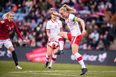 Piłkarka w białej koszulce z czerwonymi szortami kontroluje piłkę. To emocjonujący moment kobiecego futbolu.