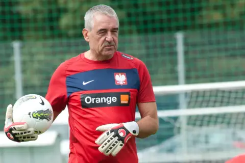 Jacek Kazimierski, trener bramkarzy, w czerwonej koszulce z orłem i logo Orange, trzyma piłkę i rękawice bramkarskie.