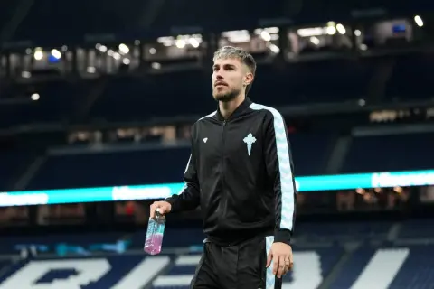 Piłkarz Oscar Mingueza w czarnej bluzie z jasnoniebieskimi pasami, trzymający butelkę napoju izotonicznego na tle stadionu.