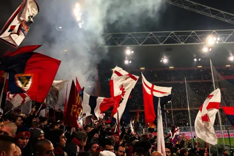 Tłum kibiców Genoa CFC z flagami na stadionie. Dym i światła tworzą atmosferę meczu.