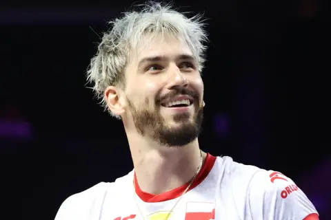Uśmiechnięty siatkarz z blond włosami, jeden z tych, którzy wygrali Ligę Mistrzów, w białej koszulce z polską flagą.
