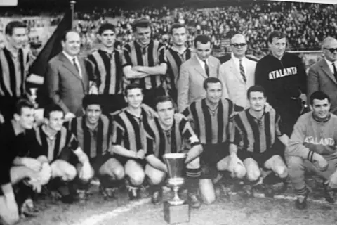 Drużyna Atalanta BC świętuje zwycięstwo z pucharem. Piłkarze w pasiatych koszulkach i sztab szkoleniowy pozują do zdjęcia.