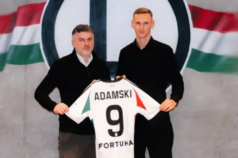 Rafał Adamski z numerem 9 na koszulce, obok niego mężczyzna, na tle flagi i logo klubu.
