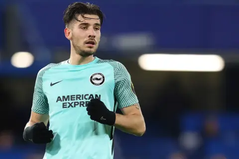 Piłkarz Jakub Moder w turkusowej koszulce Brighton & Hove Albion, z rękawiczkami na dłoniach, gotowy do gry.