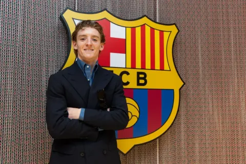 Uśmiechnięty Anthony Gordon w garniturze stoi przed herbem FC Barcelony.
