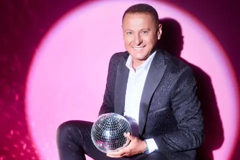 Piotr Świr Świerczewski w eleganckim garniturze trzyma błyszczącą kulę dyskotekową na tle różowego światła.