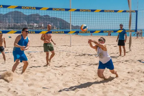 Mężczyźni grają w siatkówkę plażową. Jeden z nich zagrywa piłkę, inni czekają na boisku. To świetny przykład beach soccer Polska!