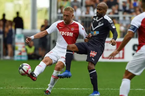 Piłkarz w białej koszulce z herbem **AS Monaco FC** walczy o piłkę z przeciwnikiem w granatowym stroju.