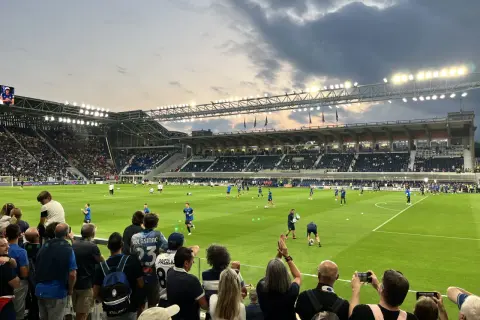 Widownia na stadionie przed meczem FC Bologna. Piłkarze rozgrzewają się na zielonej murawie, a kibice z flagami i aparatami obserwują przygotowania.
