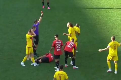 Sędzia pokazuje czerwoną kartkę w trakcie meczu Stade Rennais. Piłkarz leży na boisku, inni zawodnicy reagują.