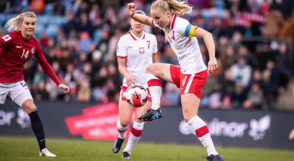 Piłkarka w białej koszulce z czerwonymi szortami kontroluje piłkę. To emocjonujący moment kobiecego futbolu.