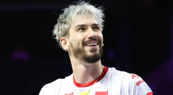 Uśmiechnięty siatkarz z blond włosami, jeden z tych, którzy wygrali Ligę Mistrzów, w białej koszulce z polską flagą.