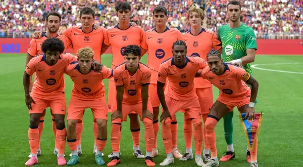 FC Barcelona - skład. Drużyna w pomarańczowych strojach pozuje do zdjęcia na boisku. W tle trybuny pełne kibiców.