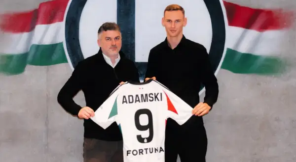Rafał Adamski z numerem 9 na koszulce, obok niego mężczyzna, na tle flagi i logo klubu.