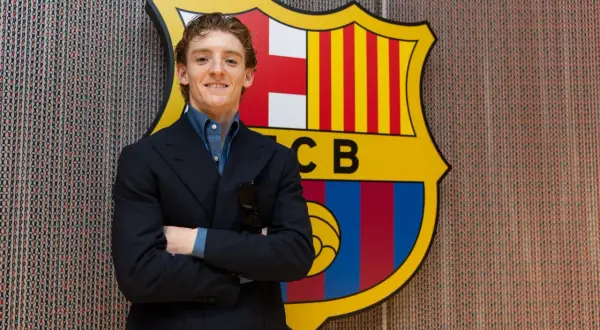 Uśmiechnięty Anthony Gordon w garniturze stoi przed herbem FC Barcelony.