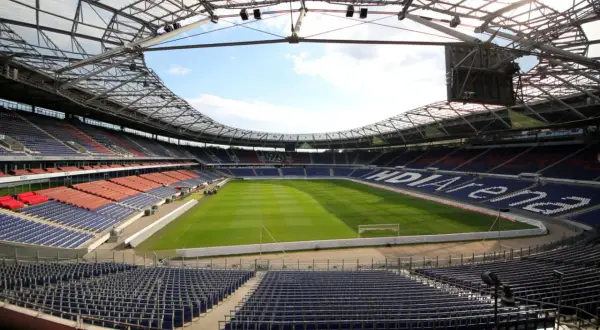 Pusta HDI Arena, stadion klubu Hannover 96, z zieloną murawą i rzędami niebieskich i czerwonych siedzeń pod przeszklonym dachem.