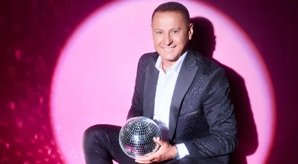 Piotr Świr Świerczewski w eleganckim garniturze trzyma błyszczącą kulę dyskotekową na tle różowego światła.