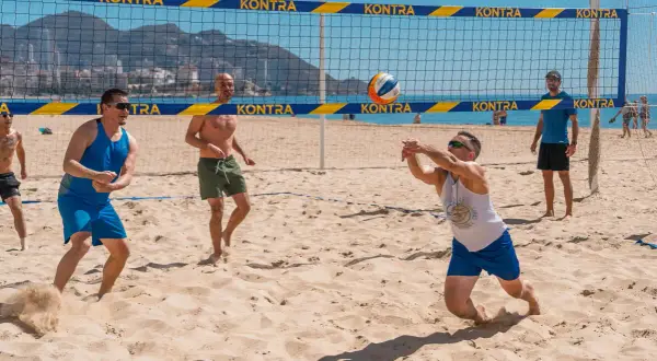 Mężczyźni grają w siatkówkę plażową. Jeden z nich zagrywa piłkę, inni czekają na boisku. To świetny przykład beach soccer Polska!