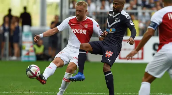 Piłkarz w białej koszulce z herbem **AS Monaco FC** walczy o piłkę z przeciwnikiem w granatowym stroju.