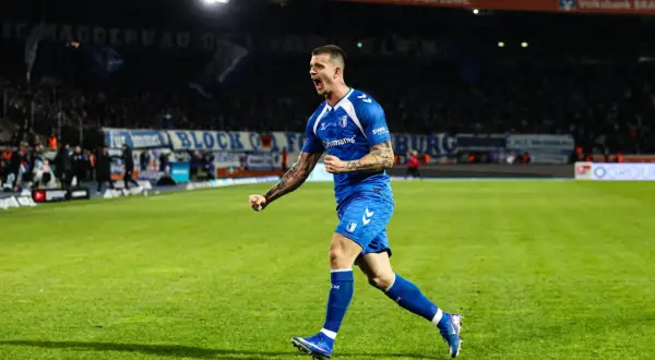 Mateusz Żukowski w niebieskim stroju świętuje bramkę na zielonej murawie stadionu, z trybunami pełnymi kibiców w tle.