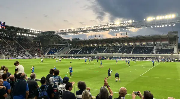 Widownia na stadionie przed meczem FC Bologna. Piłkarze rozgrzewają się na zielonej murawie, a kibice z flagami i aparatami obserwują przygotowania.