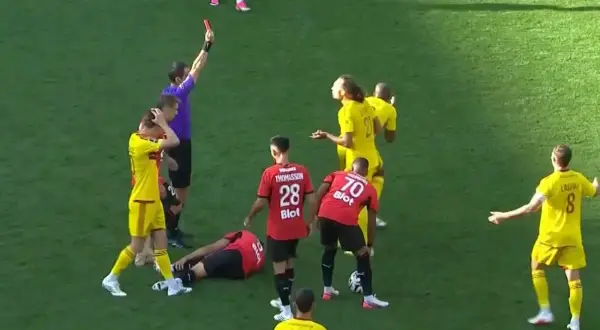 Sędzia pokazuje czerwoną kartkę w trakcie meczu Stade Rennais. Piłkarz leży na boisku, inni zawodnicy reagują.