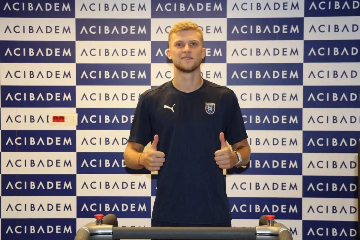 Jakub Kałuziński z podniesionymi kciukami, w granatowej koszulce z logo klubu, na tle niebiesko-białej ściany z powtarzającym się napisem.