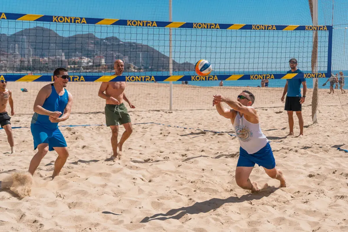 Mężczyźni grają w siatkówkę plażową. Jeden z nich zagrywa piłkę, inni czekają na boisku. To świetny przykład beach soccer Polska!