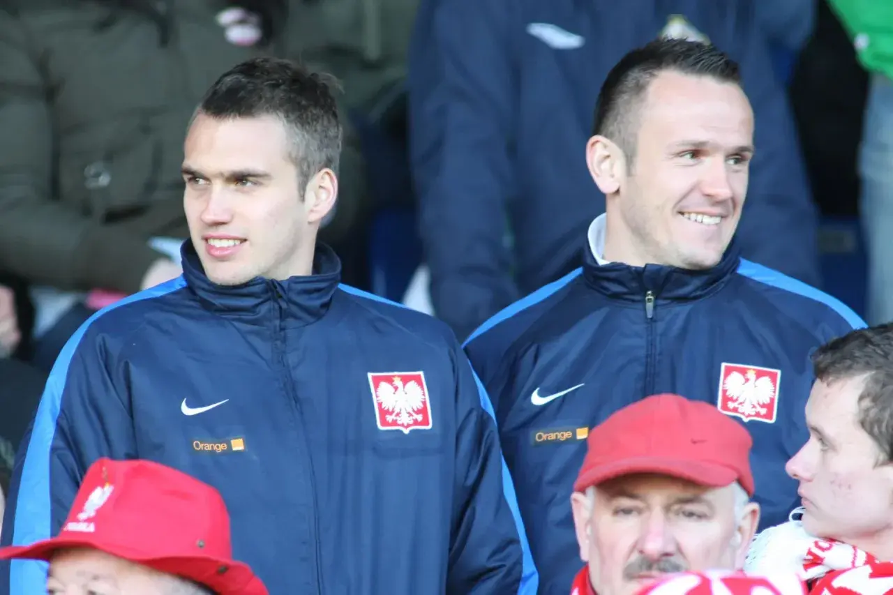 Dwóch piłkarzy w dresach reprezentacji Polski, jeden z nich to Łukasz Sosin, uśmiecha się do kibiców.