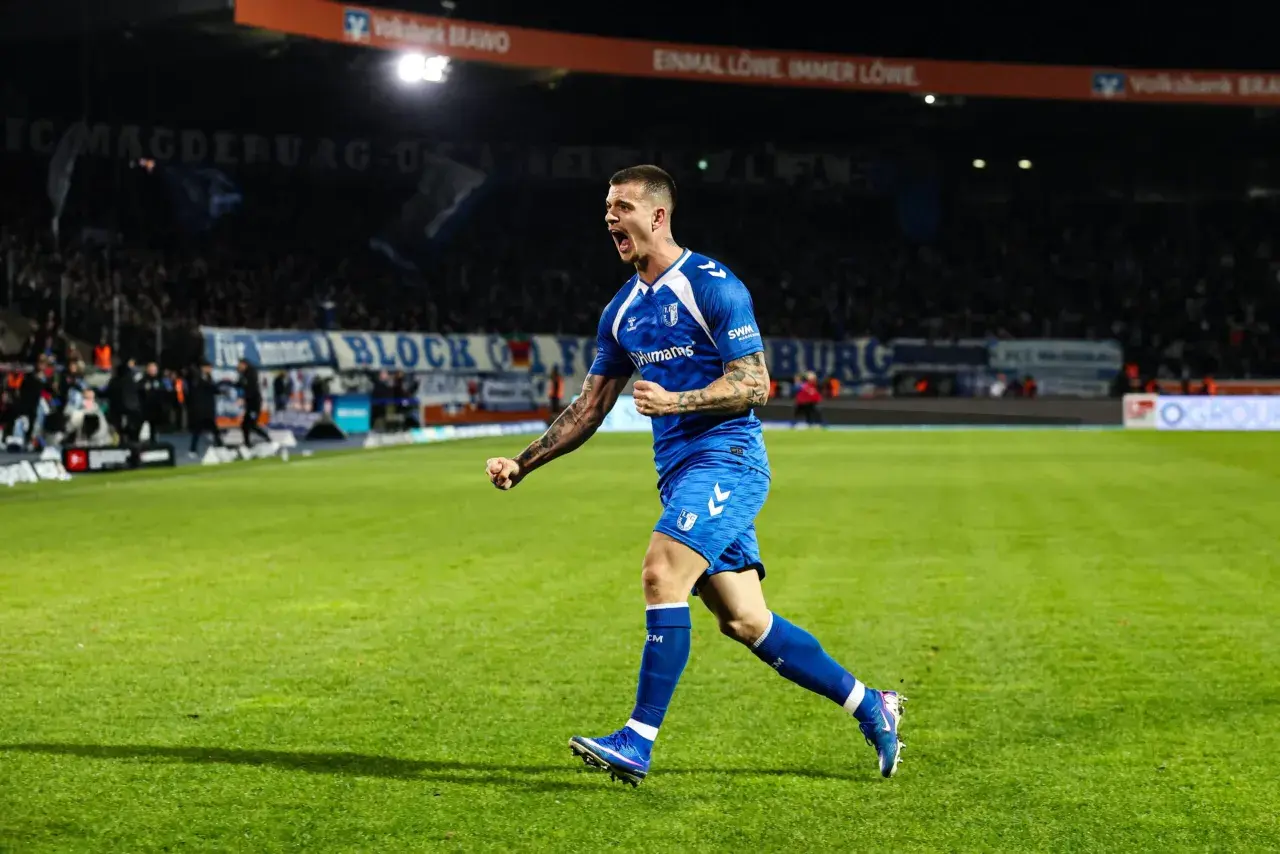 Mateusz Żukowski w niebieskim stroju świętuje bramkę na zielonej murawie stadionu, z trybunami pełnymi kibiców w tle.