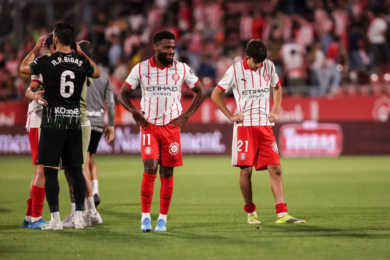 Piłkarze Girona FC po trudnych rozgrywkach. Zawodnicy w czerwono-białych strojach stoją na boisku, wyraźnie zmęczeni.