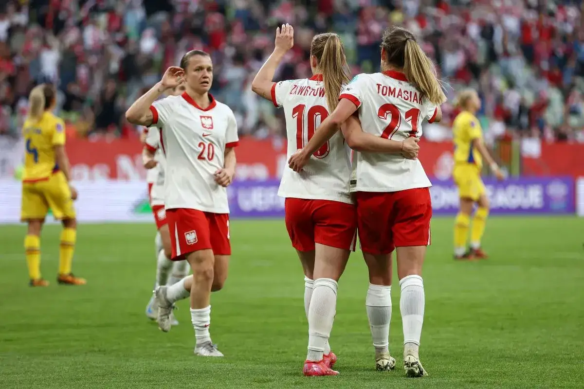 Piłkarki Polski świętują zwycięstwo nad Rumunią. Dwie zawodniczki w białych koszulkach z orłem przytulają się na boisku.
