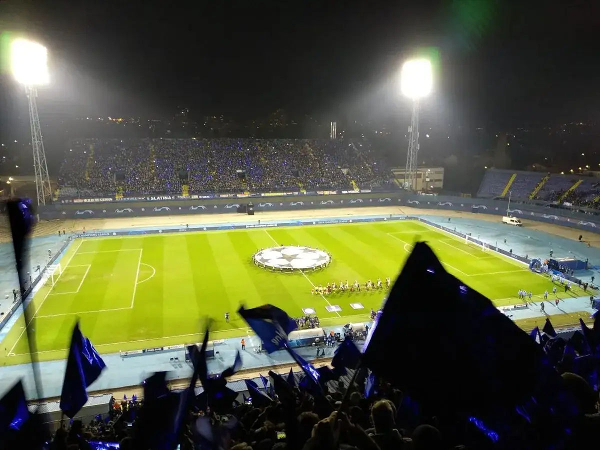 Nocny mecz na stadionie, kibice z flagami Dinamo Zagrzeb w niebieskich barwach, piłkarze na murawie.