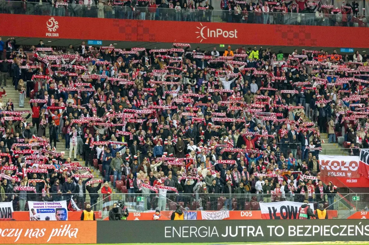 Tłum kibiców na stadionie podczas meczów ligowych, wszyscy ubrani w biało-czerwone barwy, trzymają szaliki z napisem