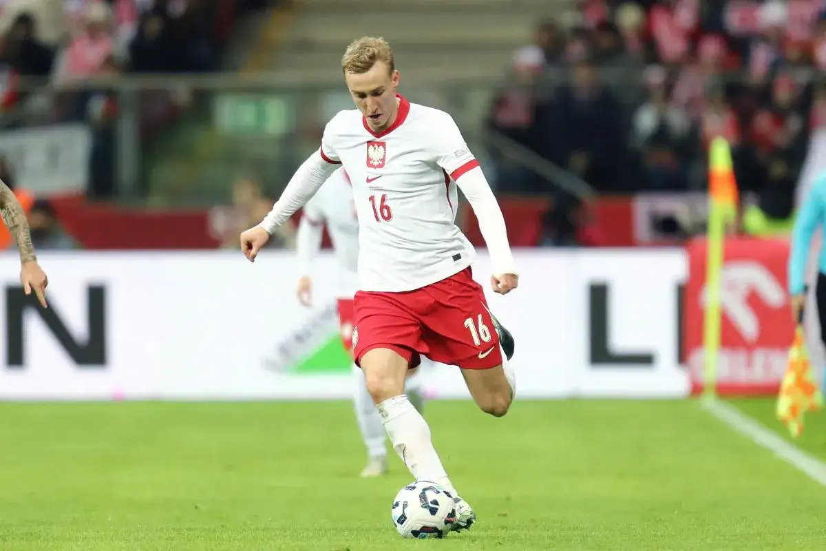 Piłkarz Adam Buksa w białej koszulce z numerem 16 i czerwonych spodenkach biegnie z piłką na boisku.