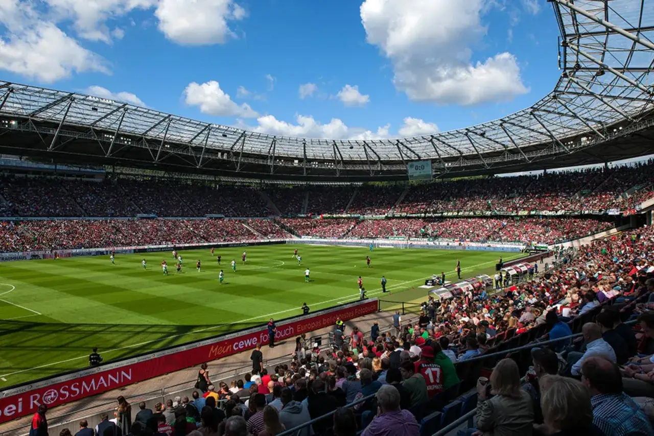 Stadion pełen kibic&oacute;w podczas meczu Hannover 96. Piłkarze na zielonej murawie, niebo z chmurami.