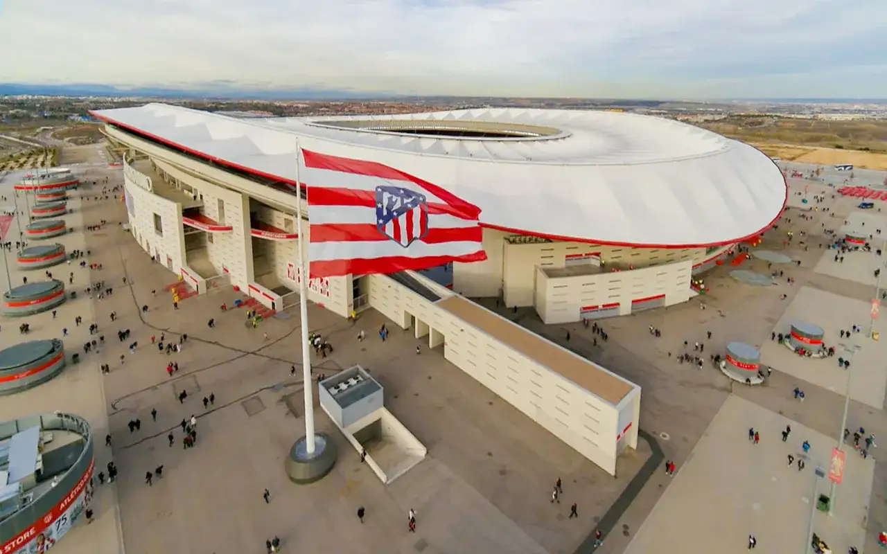 Flaga z herbem Atletico Madryt powiewa przed nowoczesnym stadionem. Wok&oacute;ł stadionu kręcą się kibice.